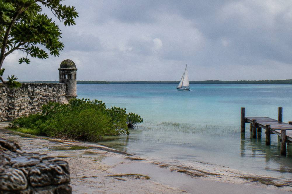 Sailboat crossing a wide stretch of Bacalar Lagoon in graduated shades of blue