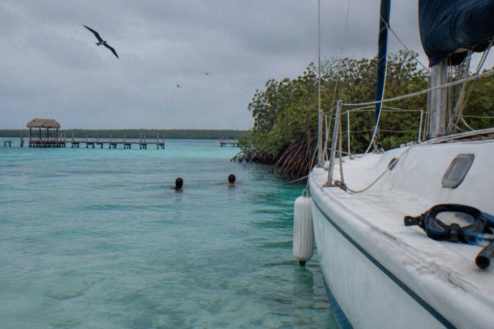 A sailboat anchored in clear shallow water with swimmers near the hull in Bacalar Lagoon