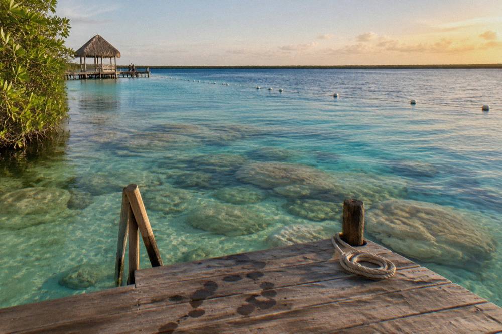 A wooden dock extending over clear green and blue lagoon water with buoys visible in the distance