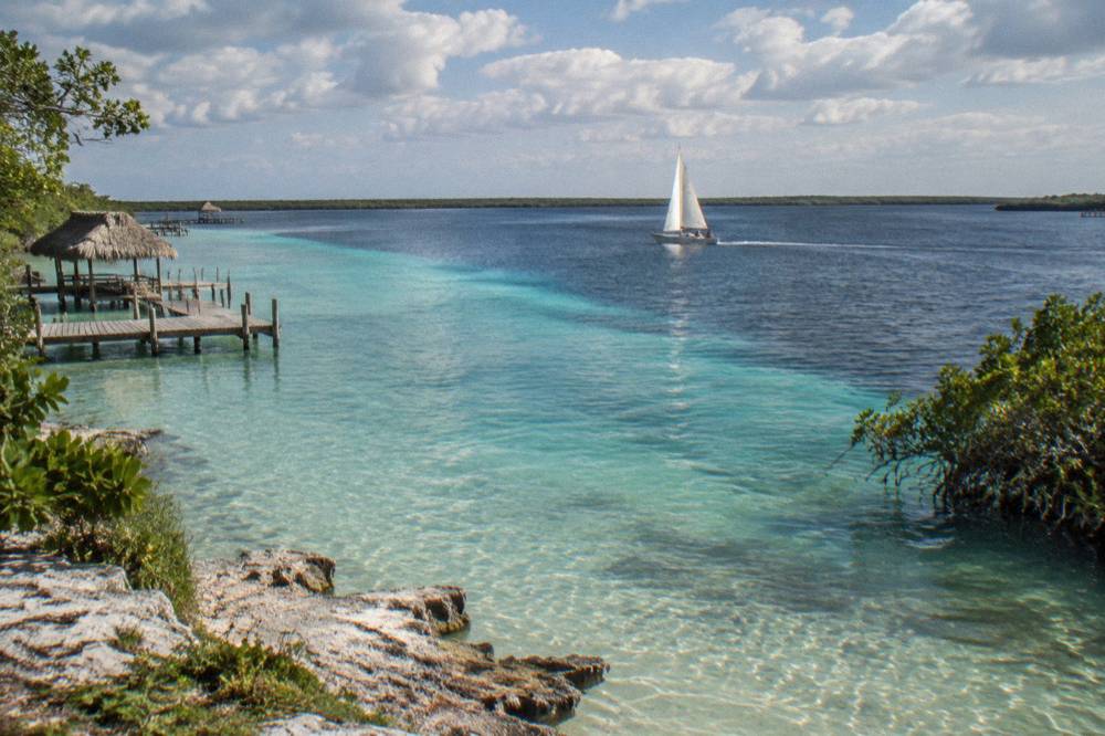 Sailboat crossing clear turquoise water in Bacalar Lagoon under scattered clouds