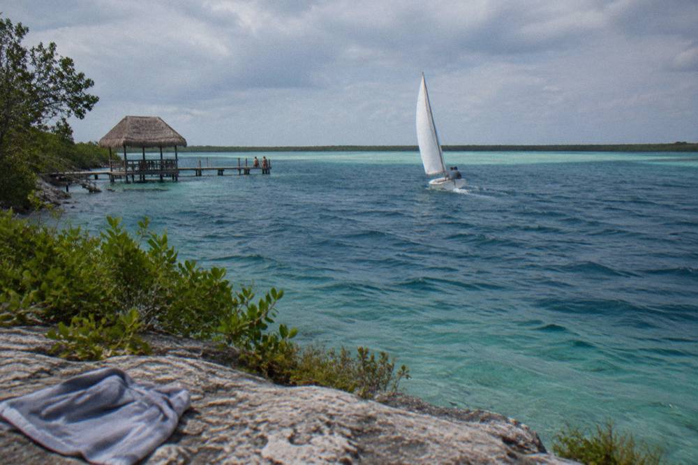 A sailboat crossing a band of deep blue water above a submerged cenote in Bacalar Lagoon