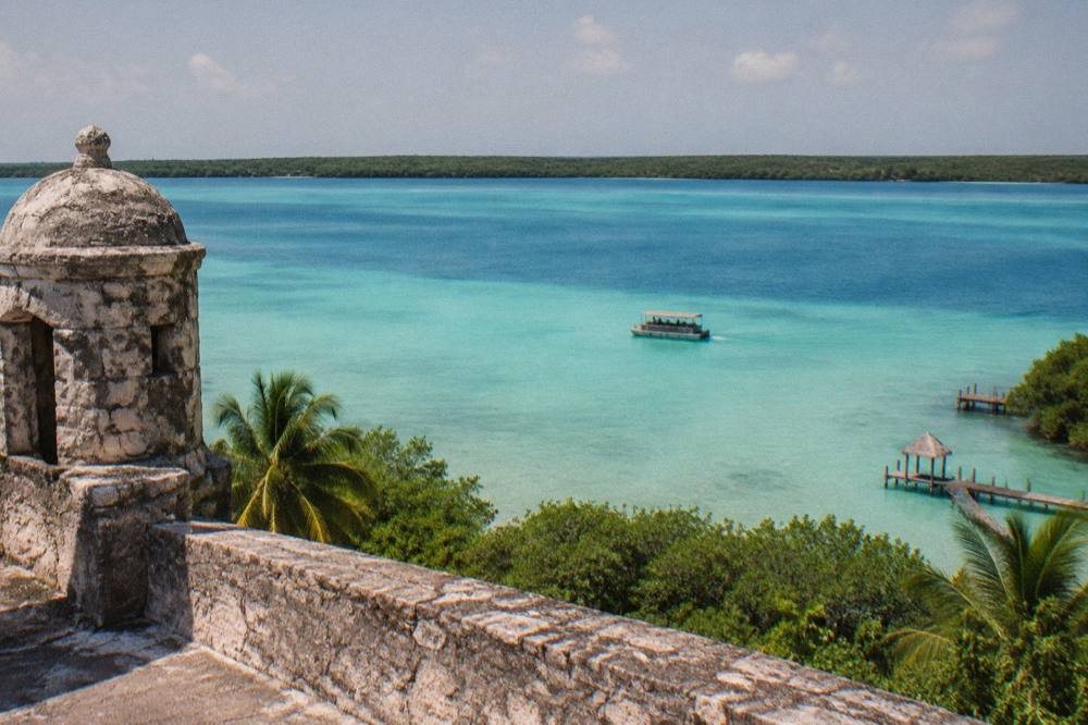 A pontoon boat crossing wide bands of blue and turquoise water on Bacalar Lagoon under midday sun
