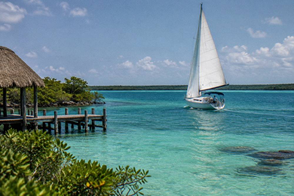 A small sailboat with a raised white sail crossing open water on Bacalar Lagoon