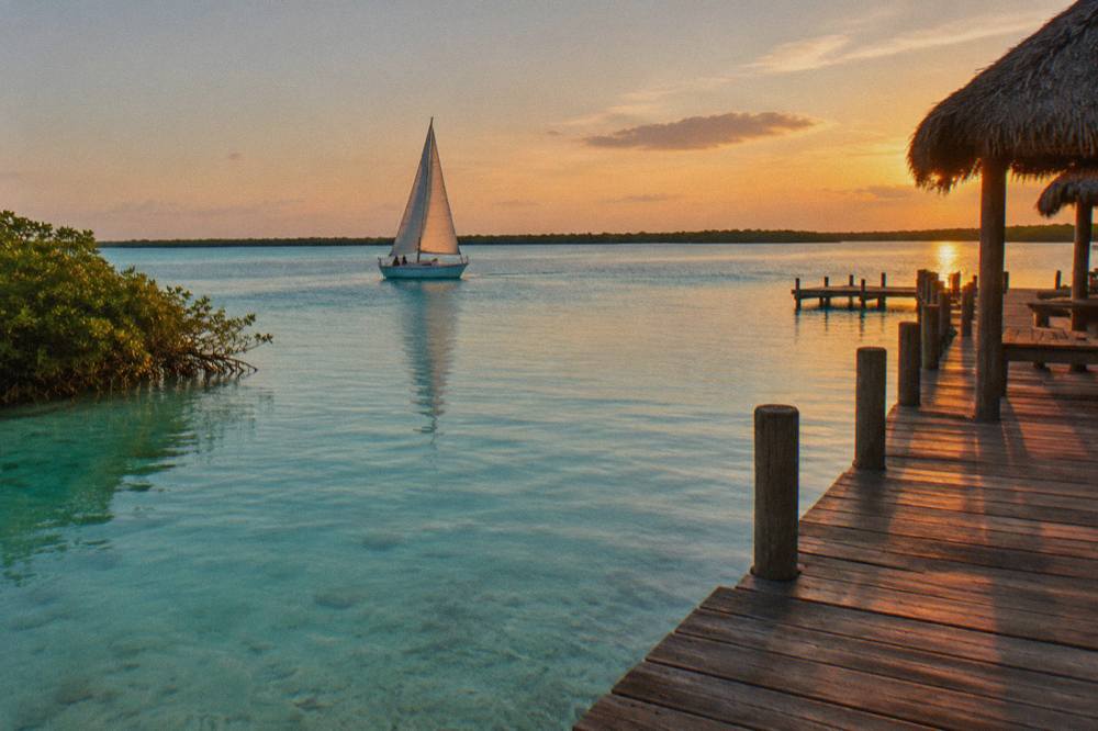 A small sailboat with its mainsail up on flat blue-green water near a wooden pier