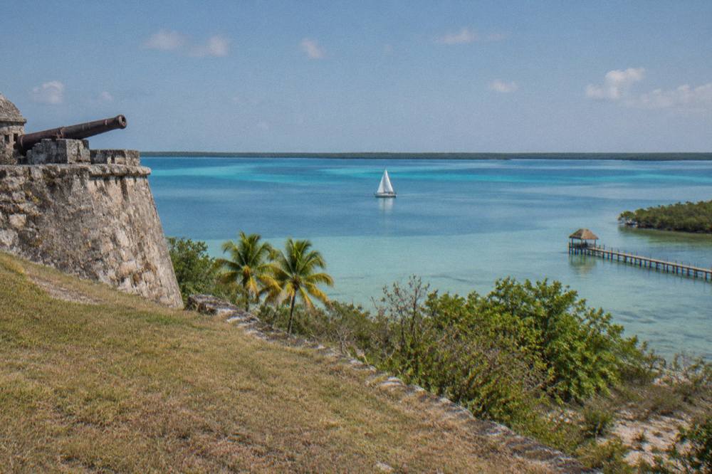 Sailboat on flat blue-green Bacalar Lagoon water under a clear morning sky