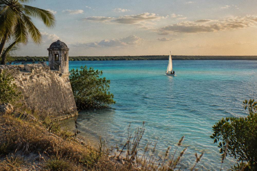 Sailboat crossing wide open turquoise lagoon water toward distant tree line