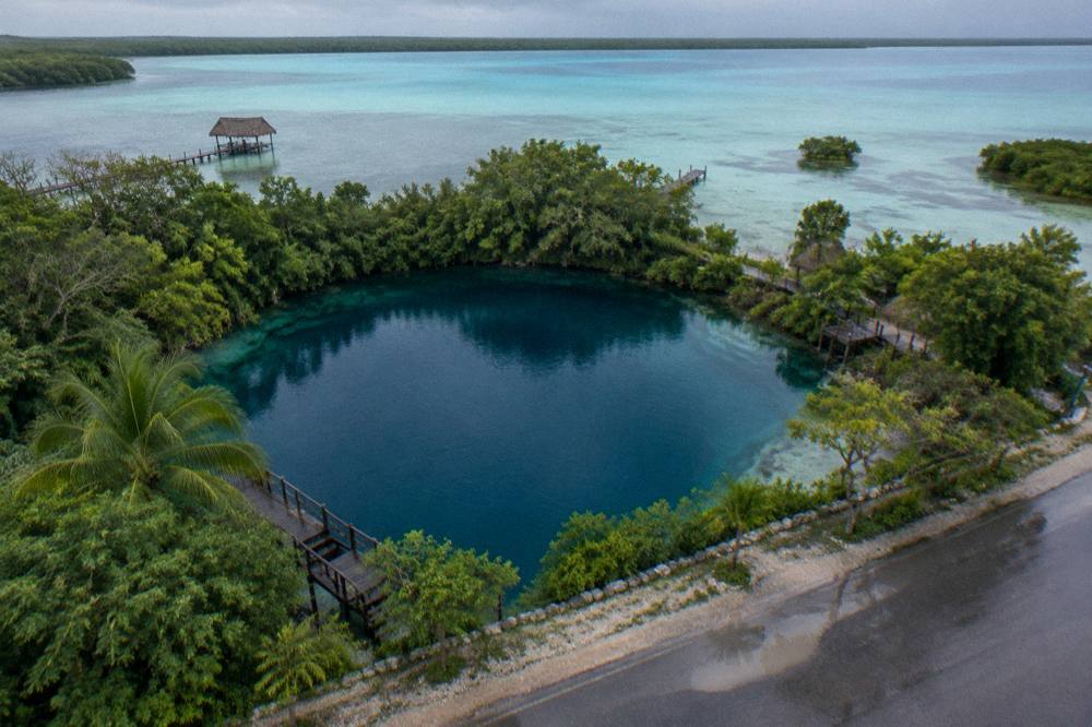 Aerial view of a round deep blue cenote beside a green shoreline road