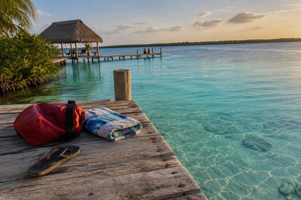 A dry bag and folded towel resting on a wooden dock above clear turquoise lagoon water