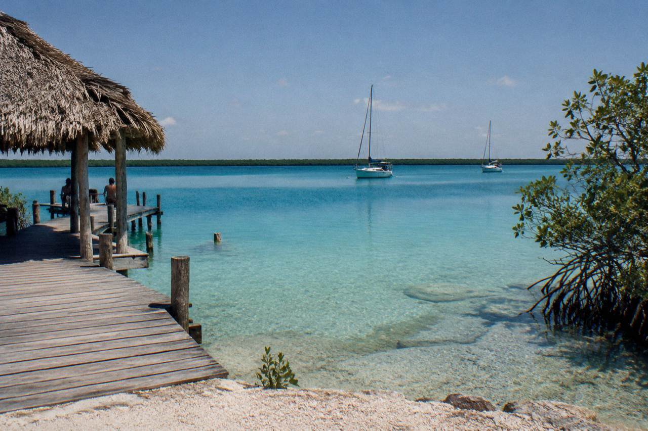 Calm turquoise water of Bacalar Lagoon at midday with a wooden dock and sailboats in the distance